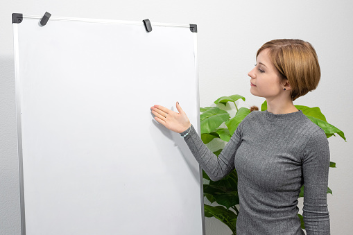 A young woman is standing in a bright room pointing at a blank flip chart. Online teacher, tutor.