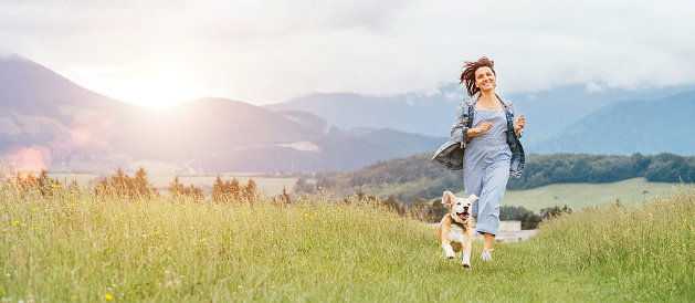 Happy smiling running beagle dog portrait with tongue out and owner female jogging by the mounting meadow grass path. Walking in nature with pets, happy healthy active people lifestyle concept image.