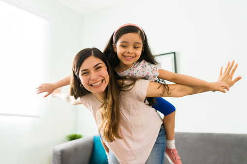 Carefree mom and little girl laughing while playing at home. Hispanic beautiful mother carrying her daughter on the back like an airplane