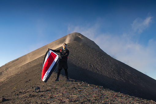 a brave and young hiker full of adrenaline poses with a flag at the base of the volcano on a sunny blue sky morning in the middle of a great adventure. Guatemala.