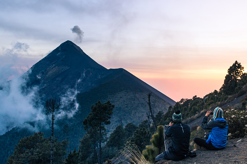 Two young hiker men contemplating the view from the Acatenango Volcano camp area where the Fuego Volcano partially covered in fog can be seen on a picturesque afternoon with dense forest. Guatemala, Acatenango.