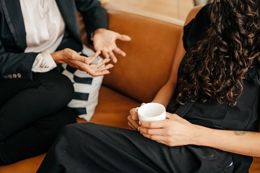 A close up shot of two female co-wokers enjoying a coffee break. Their faces are not visible. One of them has a coffee mug in her hands. They are seating on a brown sofa. Horizontal indoor daylight photo.