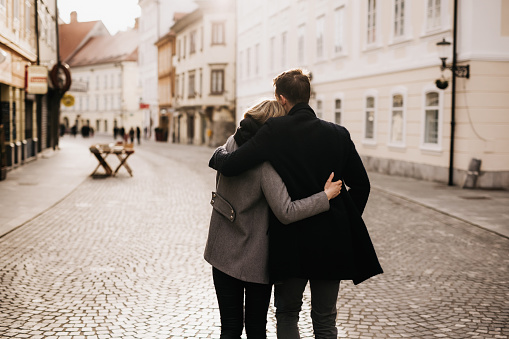 A fashionable young couple is strolling along a charming cobbled street in a European town. They are dressed in trendy winter coats. Faces are not visible. Horizontal daylight photo with copy space.