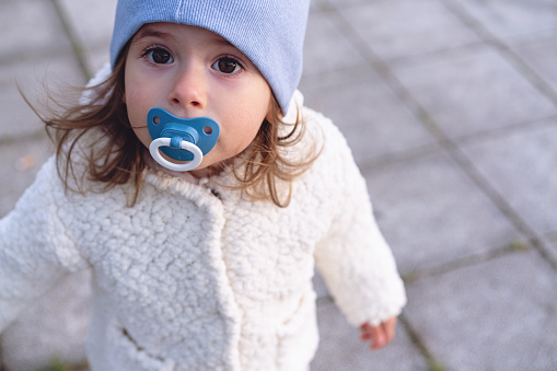 Charming and cute toddler girl in warm clothes and pacifier in her mouth exploring the day