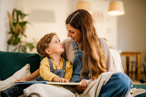 A young mother and her cute son are enjoying time together in their living room. The child is looking at his mother with the biggest eyes full of determination and curiosity. They are dressed in matching dungarees and are covered with a white blanket. Horizontal daylight indoor photo.