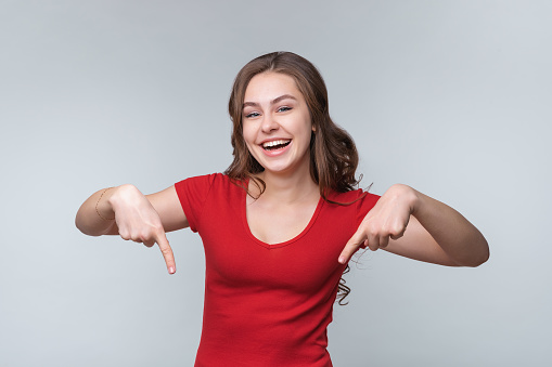 Happy young brunette woman smiling and pointing fingers down at your promo text. Cute girl student looking cheerful at camera, points hands aside at copy space logo, standing over gray background