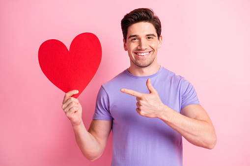 Photo of handsome cheerful person indicate finger paper heart card toothy smile isolated on pink color background.