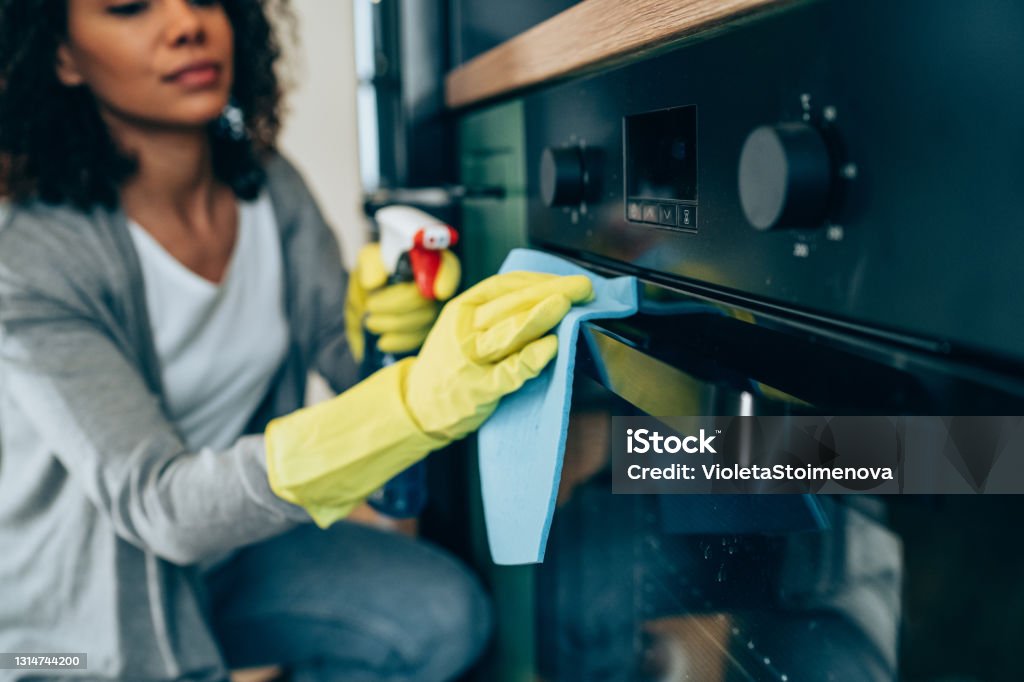 Woman wiping surface at home. Shot of young woman cleaning the outside of an oven. Woman cleaning with spray disinfectant and gloves at home. Housework Stock Photo