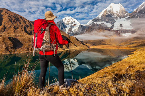 Young woman, wearing a red jacket, trekking in Peruvian Andes near Lake Carhuacocha, Mount Yerupaja on the background, Cordillera Huayhuash. Andes Mountain Range is located in South America, running north to south along the western coast of the continent.It is a continual range of highlands along the western coast of South America.The Andes extend from north to south through seven South American countries: Argentina, Bolivia, Chile, Colombia, Ecuador, Peru, and Venezuela, and is the longest continental mountain range in the world.