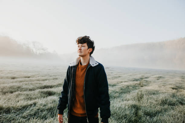 Young handsome androgynous women standing on a foggy meadow early in the morning. Short curly brown hair. looking away.
