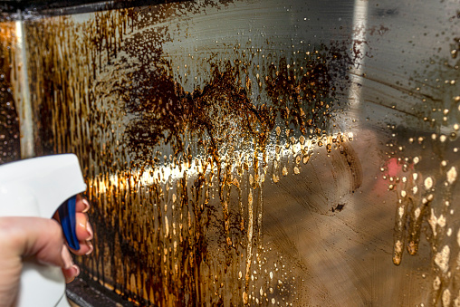 A woman spraying a corner glass pane covered in soot in a modern fireplace with a closed combustion chamber standing in the living room.