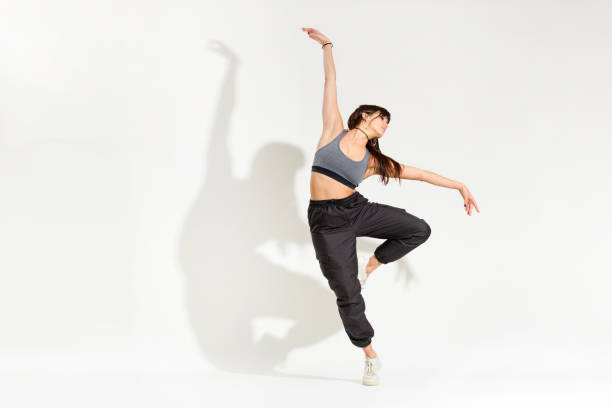 Graceful young woman dancer performing a classic dance pose Graceful young woman dancer in a hip hop outfit performing a classic dance pose with outstretched arms balancing on one leg with artistic shadow on a white studio background ballerina shadow stock pictures, royalty-free photos & images