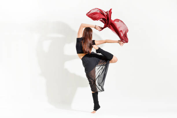 Woman dancer performing an alternative arabesque dance pose gracefully flicking red fabric above her head in a rear view over white with shadow and copyspace
