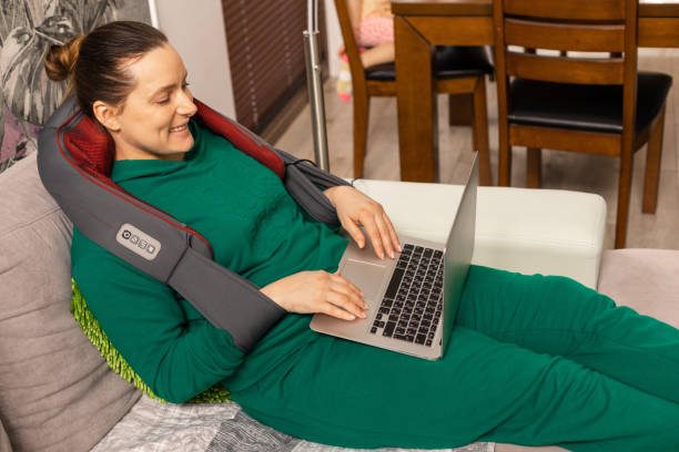 mulher trabalha em casa usando laptop e gosta de massagem no ombro e pescoço com aparelho elétrico. osteocondrose. sorri e recebe emoções positivas. equipamento elétrico - massaging alternative medicine headache women - fotografias e filmes do acervo