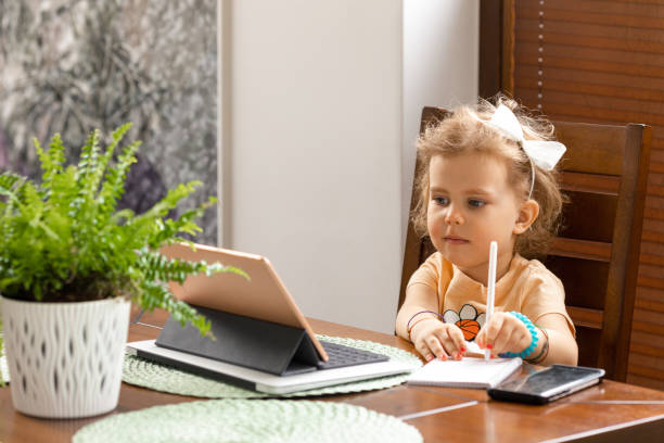 belle petite fille de 3 ans avec les cheveux bouclés est assis à table et étudie en ligne avec la tablette à la maison. concept d’éducation de la petite enfance - child caucasian little girls 3 4 years photos et images de collection