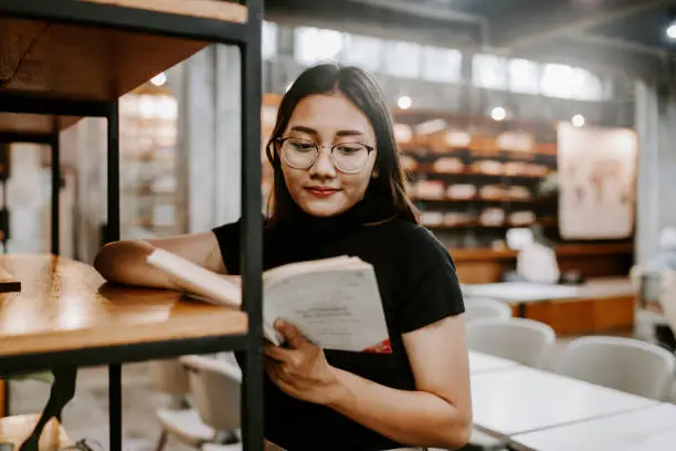 Photo of Young female student choosing book from shelf in library at community college