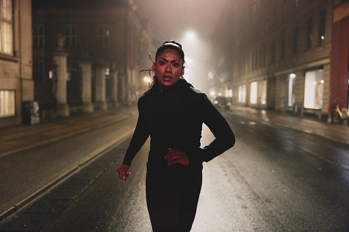 Fit young Indian woman in sportswear running on a city street at night illuminated by street lights