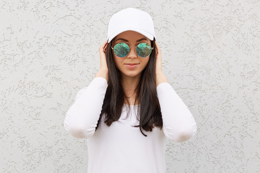 Serious confident woman wearing trendy sunglasses, baseball cap and white shirt, looks at camera, keeping hands on her head, posing isolated over light background outdoor.