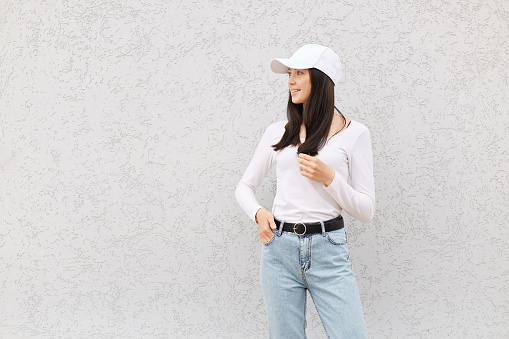 Beautiful European female wearing white shirt, jeans and baseball cap, looking away, expressing positive emotions, copy space for promotional text.