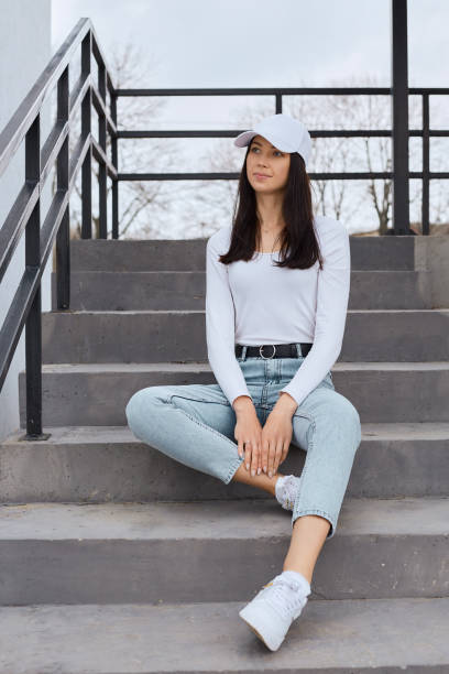 Young attractive dark haired female sitting on stairs and looking away with pensive dreamy facial expression, stylish woman wearing jeans, white shirt and baseball cap posing outdoors. Young attractive dark haired female sitting on stairs and looking away with pensive dreamy facial expression, stylish woman wearing jeans, white shirt and baseball cap posing outdoors. woman wearing baseball cap stock pictures, royalty-free photos & images