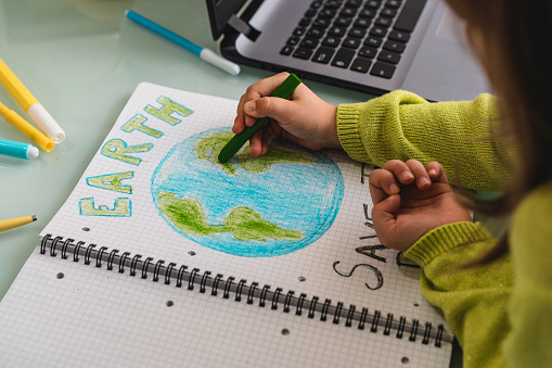 Wide angle view of child girl draws planet earth with wax colors on school notebook for Earth day - Little activist girl writes the message Save the Planet - Protection of environment, global warming