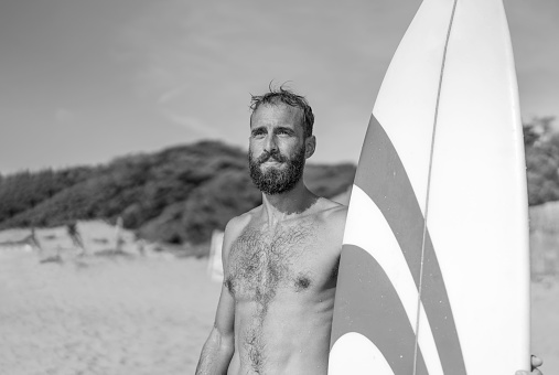 Surfer holding his surfboard on the beach - Hipster man standing on the beach and waiting big waves for surfing - Fit bearded man training with surfboard to sea - Lifestyle and freedom concept