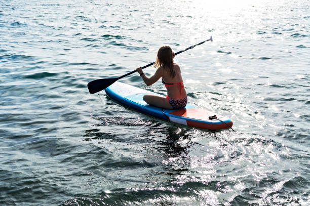giovane donna da dietro seduta e pagaiata su una tavola da paddle stand up sul mare blu, attività all'aperto - paddleboard oar women lake foto e immagini stock