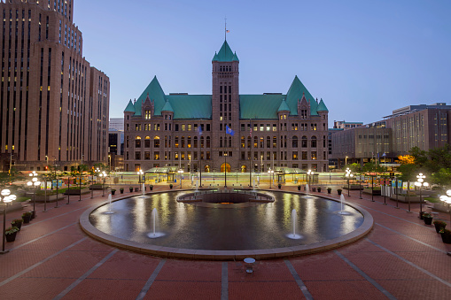 A Wide Angle Long Exposure Shot of Minneapolis' City Hall Building, Lightrail Train Station with Trains Passing, and Reflections in a Water Fountain