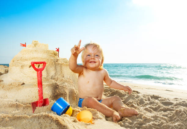 Happy cute little toddler boy sit near sand castle point finger up and smile