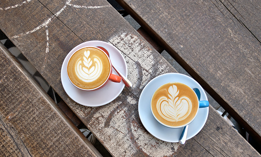 A top-view of a two cups of cappuccino with latte art at the midpoint of a wooden table