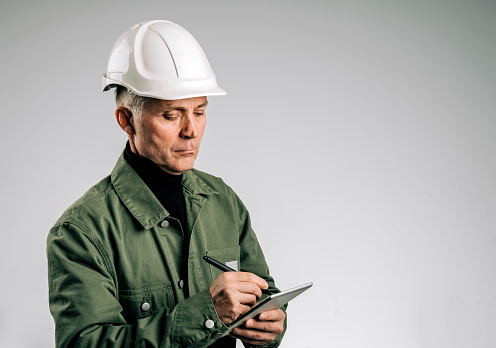 Senior man wearing hardhat making notes in digital tablet on white background. Portrait of construction worker using tablet pc.