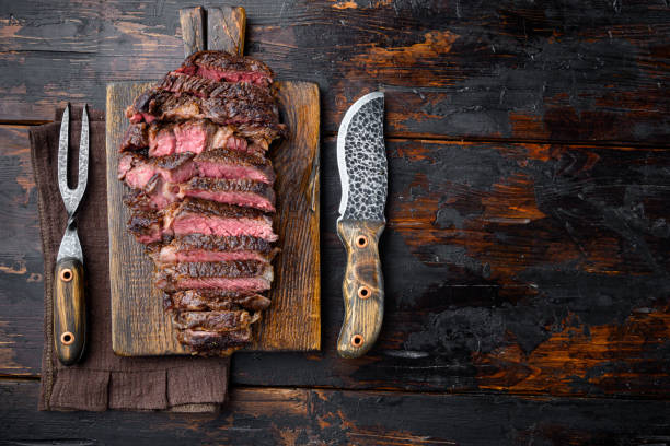 Grilled or fried and sliced marbled meat steak rib eye set, on wooden serving board, with meat knife and fork, on old dark wooden table background, top view flat lay, with copy space for text ojo de costilla de carne de marmolado a la parrilla o frito y en rodajas, en tabla de servir de madera, con cuchillo de carne y tenedor, sobre el antiguo fondo de la mesa de madera oscura, la vista superior plana yacía, con espacio de copia para el texto