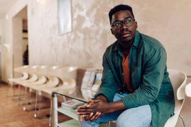 Worried African American young man waiting for his medical exam at the clinic African American patient sitting in the waiting room at the hospital and worrying for his health. waiting room stock pictures, royalty-free photos & images