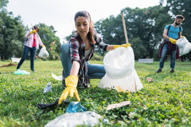 Young woman picking up litter thrown away in the park Three students cleaning local park, collecting litter in bags littering stock pictures, royalty-free photos & images