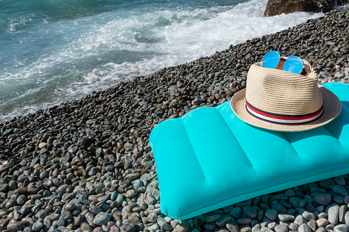 Blue inflatable swimming pillow, sunglasses on sun hat lie on  seaside near sea with waves. Travelling concept, planning summer sea vacation.