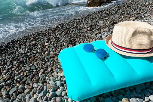 Bright cian inflatable swimming pillow, mirror sunglasses and sun hat lie on stone seaside near sea. Travelling concept, planning summer vacation on sea.