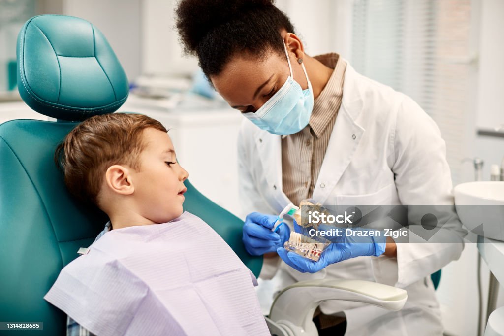 African American dentist teaching small boy how to brush teeth properly during dental appointment. Small boy sitting in dentist's chair and learning how to brush teeth properly. Dentist Stock Photo