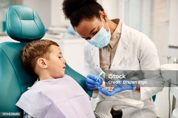 African American Dentist Teaching Small Boy How To Brush Teeth Properly During Dental Appointment Stock Photo - Download Image Now