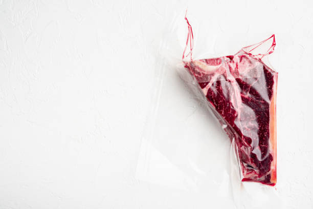 istock Raw meat T bone steak in vacuum plastic bag for sous vide cooking, on white stone  background, top view flat lay, with copy space for text 1314829481