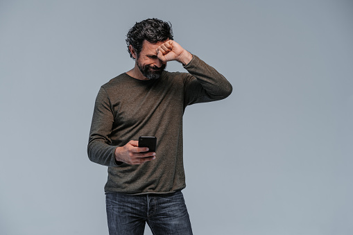 Studio shot of a repenting looking mature man with casual clothings and with his mobile phone one hand and touching his head making a fist with the other one standing on grey background. Part of a series with similar poses and different facial expressions.