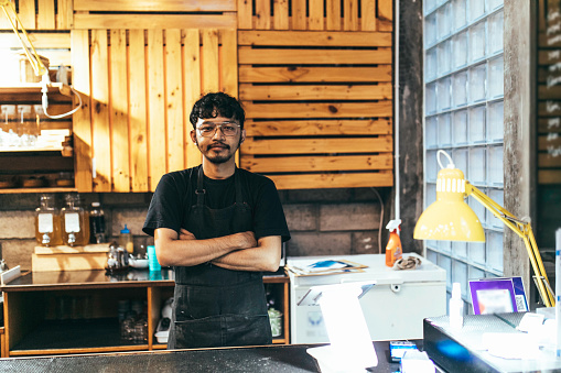 Portrait of confident barista in cafe. Young man is standing with arms crossed in coffee shop. He is wearing denim apron.