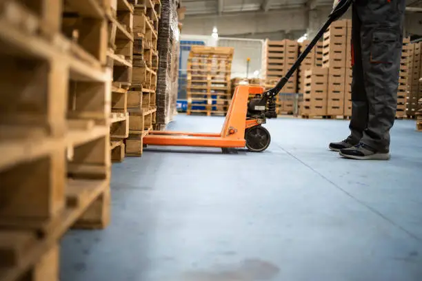 Working at warehouse. Low angle view of unrecognizable worker lifting palette with manual forklift.