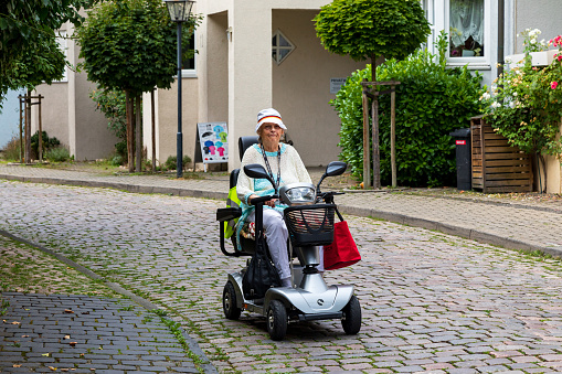 An elderly satisfied lady rides an electric scooter along the street of the old German city. Image of active old age, a taste for life, vitality in old age.. Bad Wimpfen, Germany - August 8, 2019