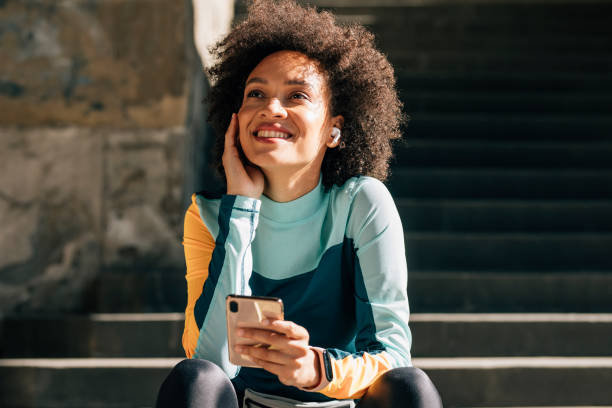 jeune athlète femelle de sourire s’asseyant sur les escaliers et écoutant la musique sur son smartphone - exercising motivation looking up african descent photos et images de collection