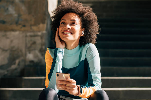 uśmiechnięta młoda zawodniczka siedząca na schodach i słuchając muzyki na swoim smartfonie - exercising motivation looking up african descent zdjęcia i obrazy z banku zdjęć