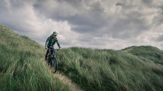 Man with bike in sand dunes with grass at coast in Denmark, dark clouds in dramatic sky