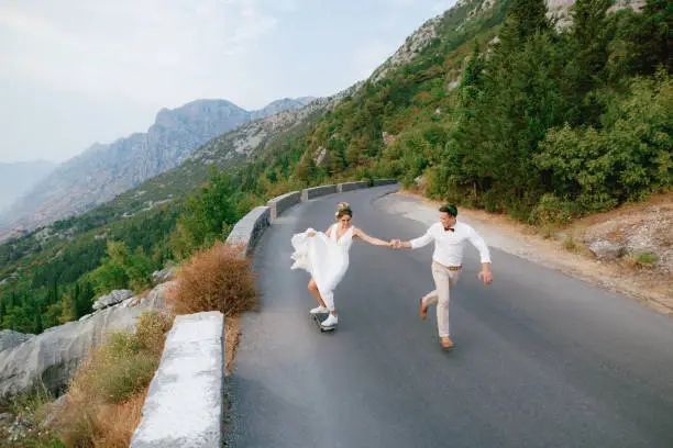 Photo of The bride rolls on a skateboard and holds the hand of the groom, who runs alongside along the highway in the mountains