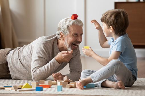 Overjoyed older Caucasian grandfather engaged in funny playful game activity with little preschooler grandson at home. Smiling mature granddad play build with bricks blocks with small boy child.