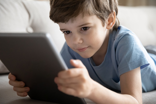 Close up of smart little 8s Caucasian boy child relax on sofa at home look at tablet screen play online game. Happy small kid use pad talk on webcam digital call, study distant. Virtual event concept.