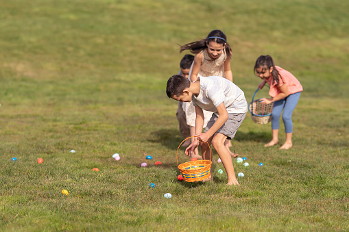 A multi-ethnic group of elementary age kids race barefoot through the grass while participating in an Easter Egg Hunt in a suburban neighborhood park. The friends are having fun celebrating the holiday together on a warm, sunny day.